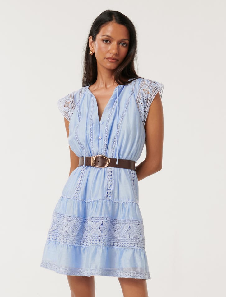 A woman wearing a light blue mini dress with lace-trimmed cap sleeves, V-neckline with tie, a belt at the waist, and a tiered, ruffled skirt.