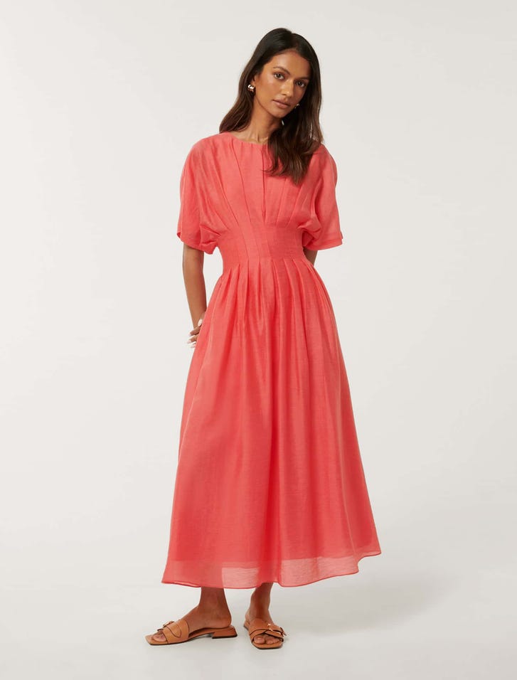 A woman wearing a coral pink midi dress with puff sleeves, a round neckline, and a full ankle-length skirt, standing against a plain background.