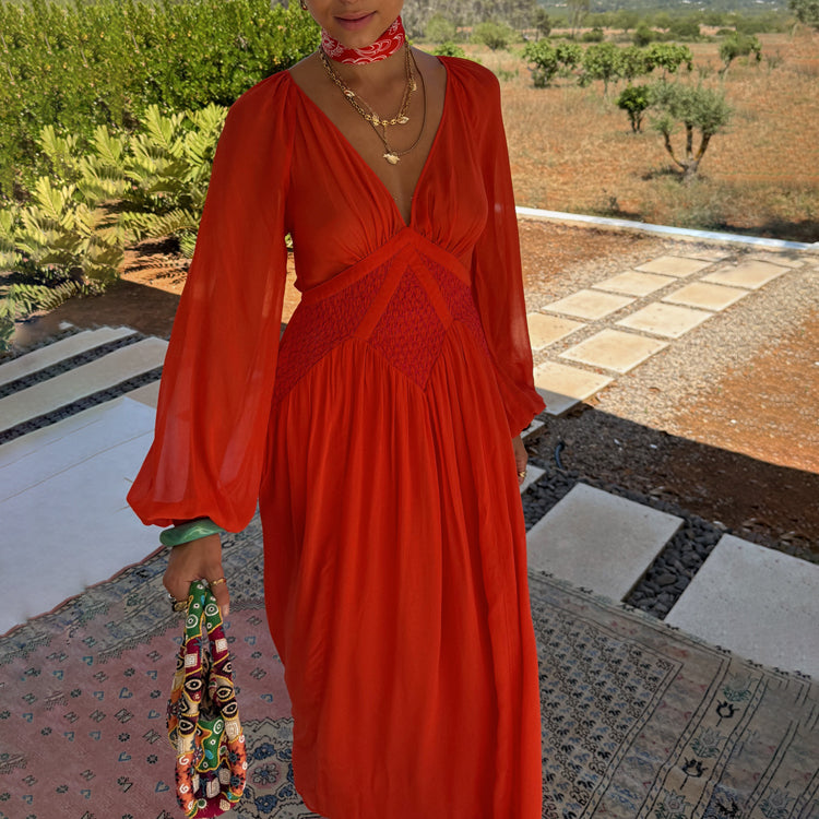 A woman wearing a red and pink dress with blouson sleeves, shirred waist, and pink smocking detail, standing outdoors.