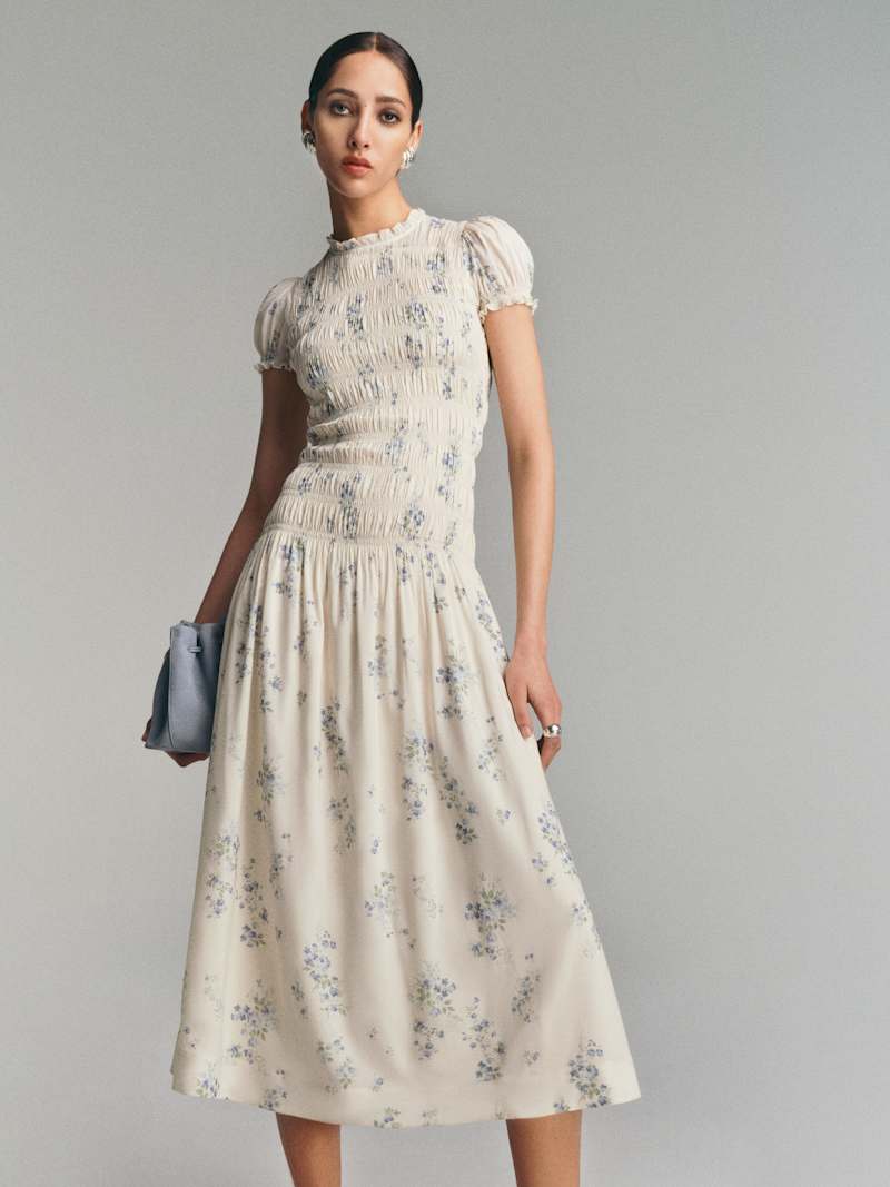 A woman wearing a light cream midi dress with floral pattern, smocked bodice, drop waist, and puff sleeves, holding a small clutch.