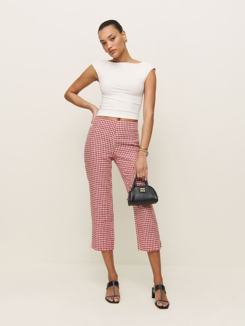 A woman wearing red and white gingham linen capri pants with a split hem, paired with a white sleeveless top, black high-heeled sandals, and holding a small black handbag.