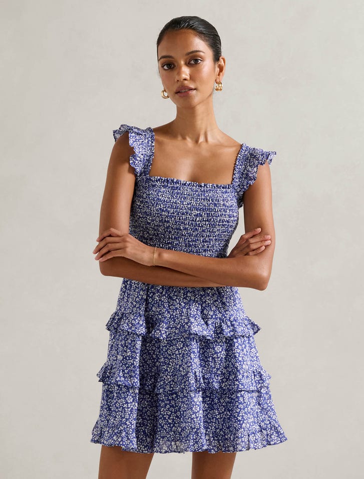 A woman wearing a blue floral mini dress with ruffled shoulder straps, square neckline, and tiered skirt.