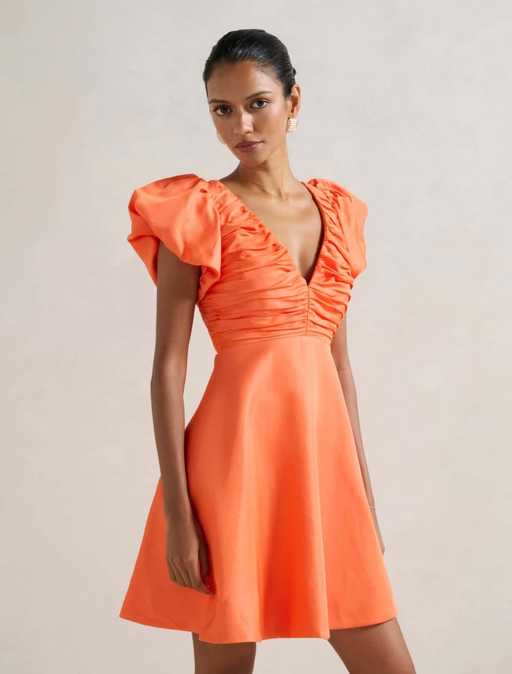 A woman wearing an orange mini dress with puffed short sleeves, a V-neckline, and a ruched bodice, standing against a plain background.