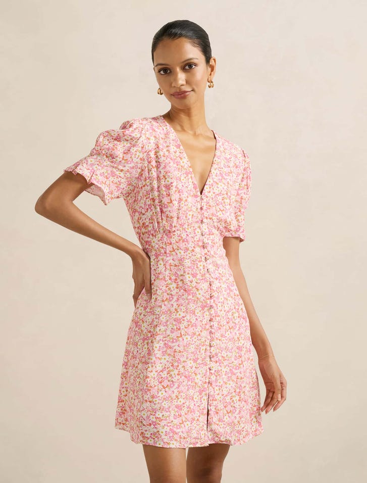 A woman wearing a pink floral petite sun dress with a V-neckline, short puffed sleeves, and a button-down front.