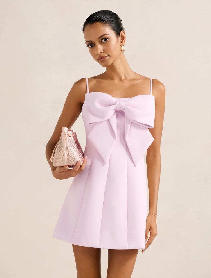 A woman wearing a light pink mini dress with a large bow at the front, square neckline, and thin shoulder straps, holding a small pink handbag.