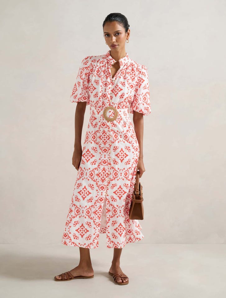 A woman wearing a white midi dress with coral embroidery, puff sleeves, stand collar, gold buttons, and a statement belt, paired with brown sandals and a handbag.