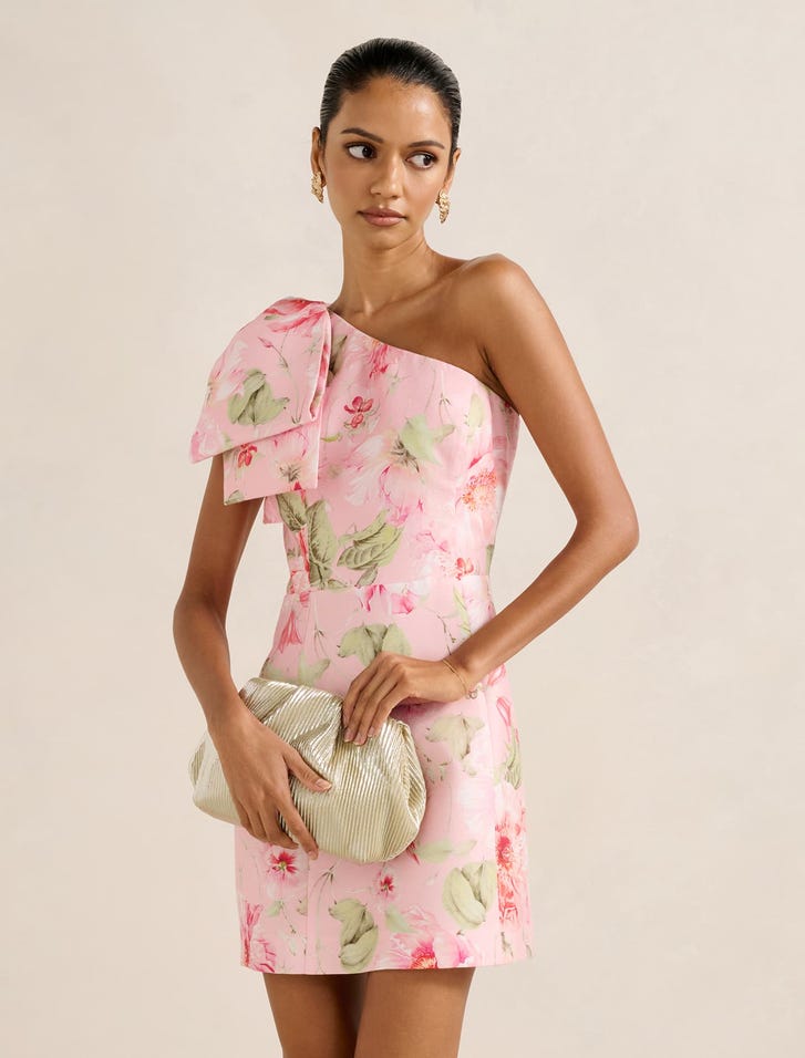 A woman wearing a pink floral one-shoulder mini dress with a large bow on the shoulder, holding a beige clutch, with her hair tied back.