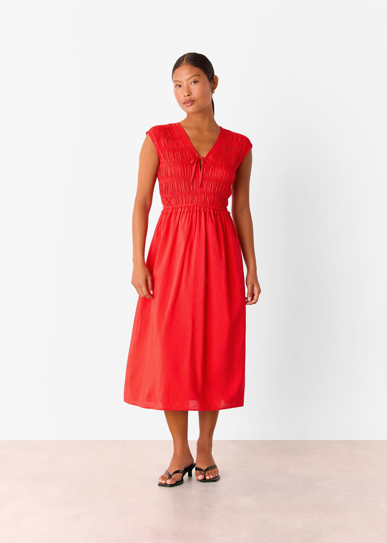 A woman wearing a red petite shirred bodice V-neck dress with a flowy hem and knot detail at the front.