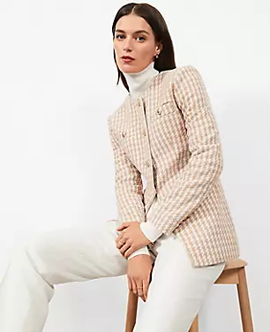 A petite woman wearing a beige and cream houndstooth tweed jacket with a button front, long sleeves, welt pockets, and a crew neck.