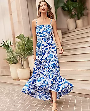 A woman wearing a blue and white tiled maxi dress with spaghetti straps, tiered design, and hi-low hem, walking outdoors near stairs and potted plants.