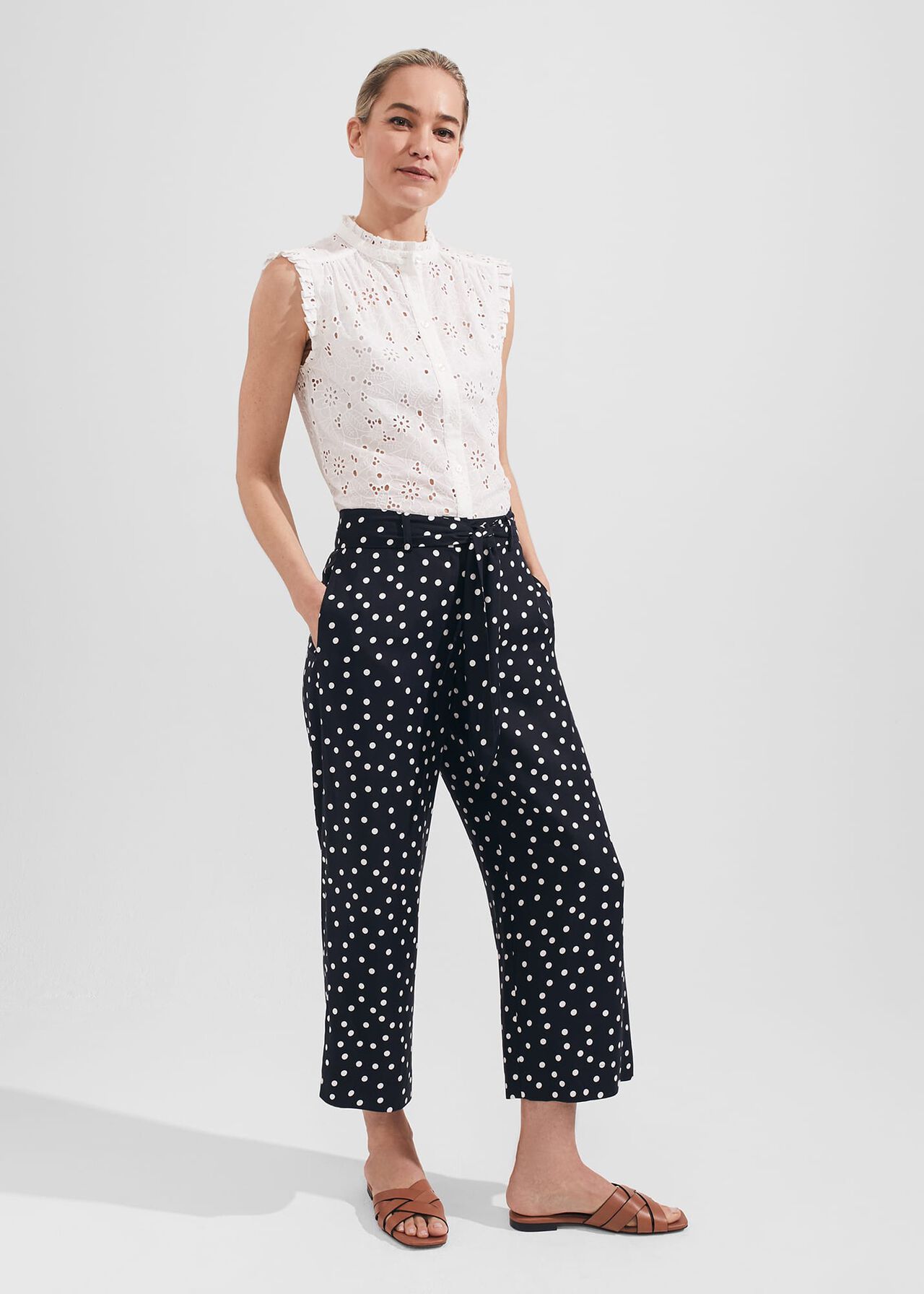 A woman wearing navy cropped trousers with white polka dots, a white sleeveless top with eyelet details, and brown sandals.