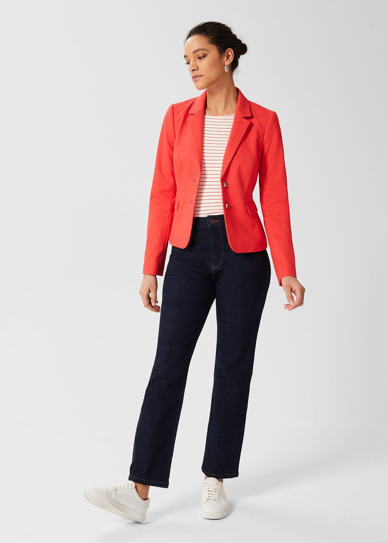 A woman wearing a coral petite Hackness jacket with a contrast Melton undercollar, faux horn buttons, and front flap pockets, paired with dark jeans and white sneakers.
