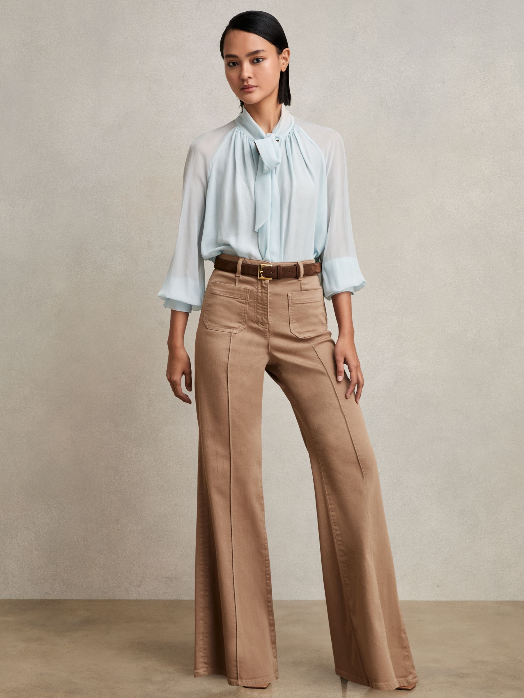 A woman wearing taupe patch-pocket flared jeans with a high waist, front pockets, and a button and zip closure, paired with a light blue blouse.