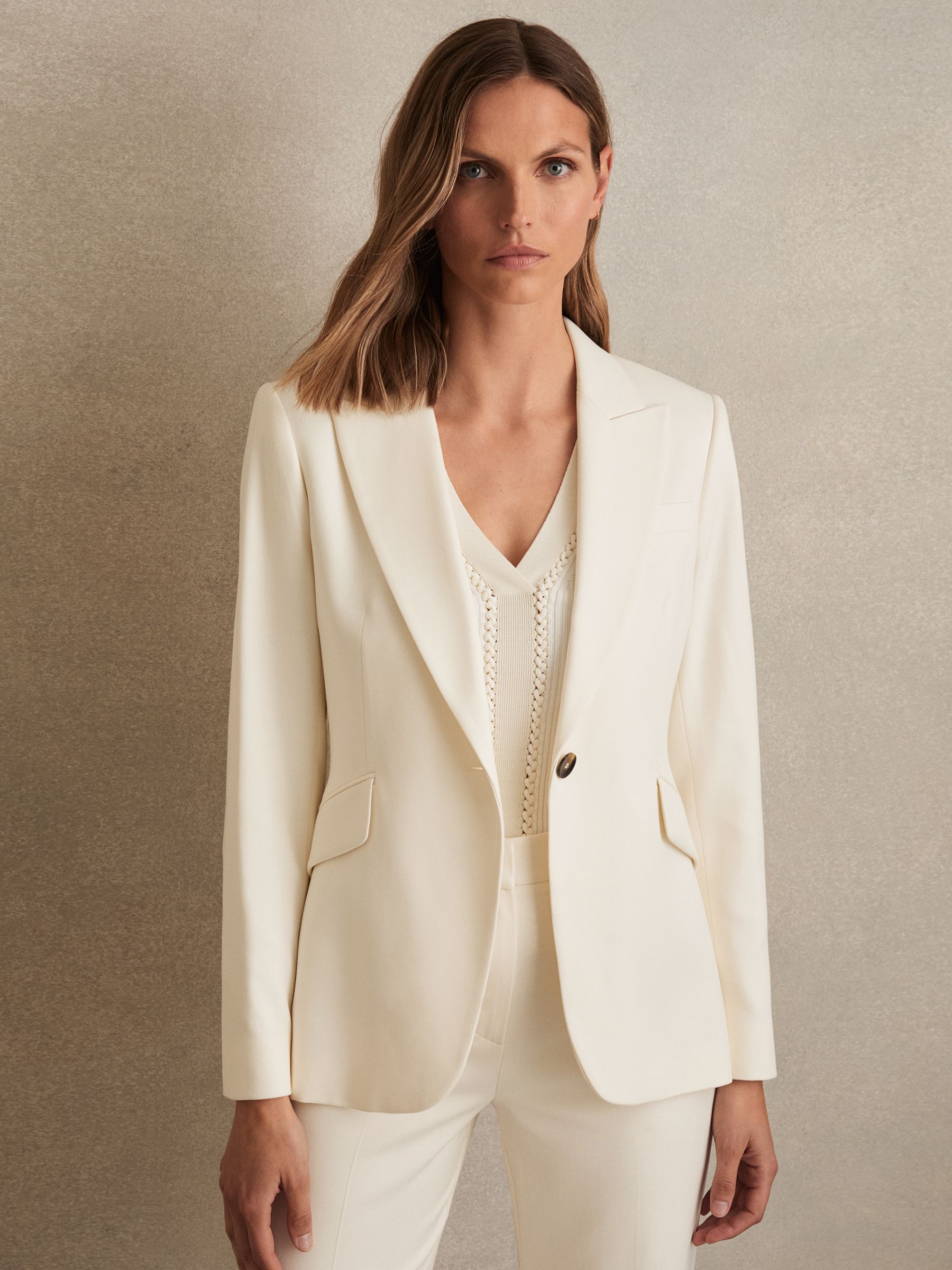 A woman wearing a cream tailored blazer with peak lapels, front flap pockets, and a single button, styled with a matching cream top underneath.