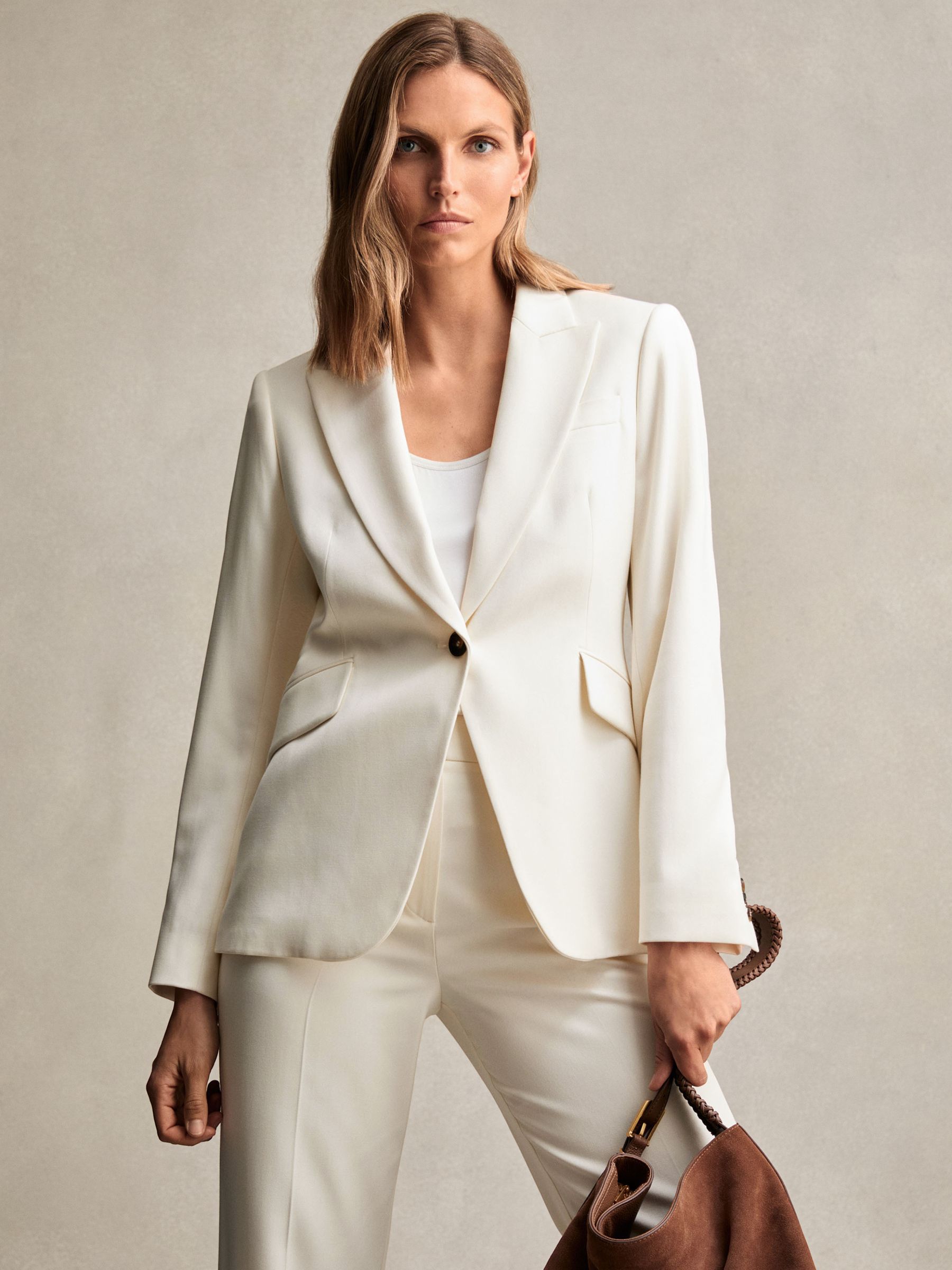 A woman wearing a cream tailored blazer with peak lapels, single-breasted closure, and flap pockets.