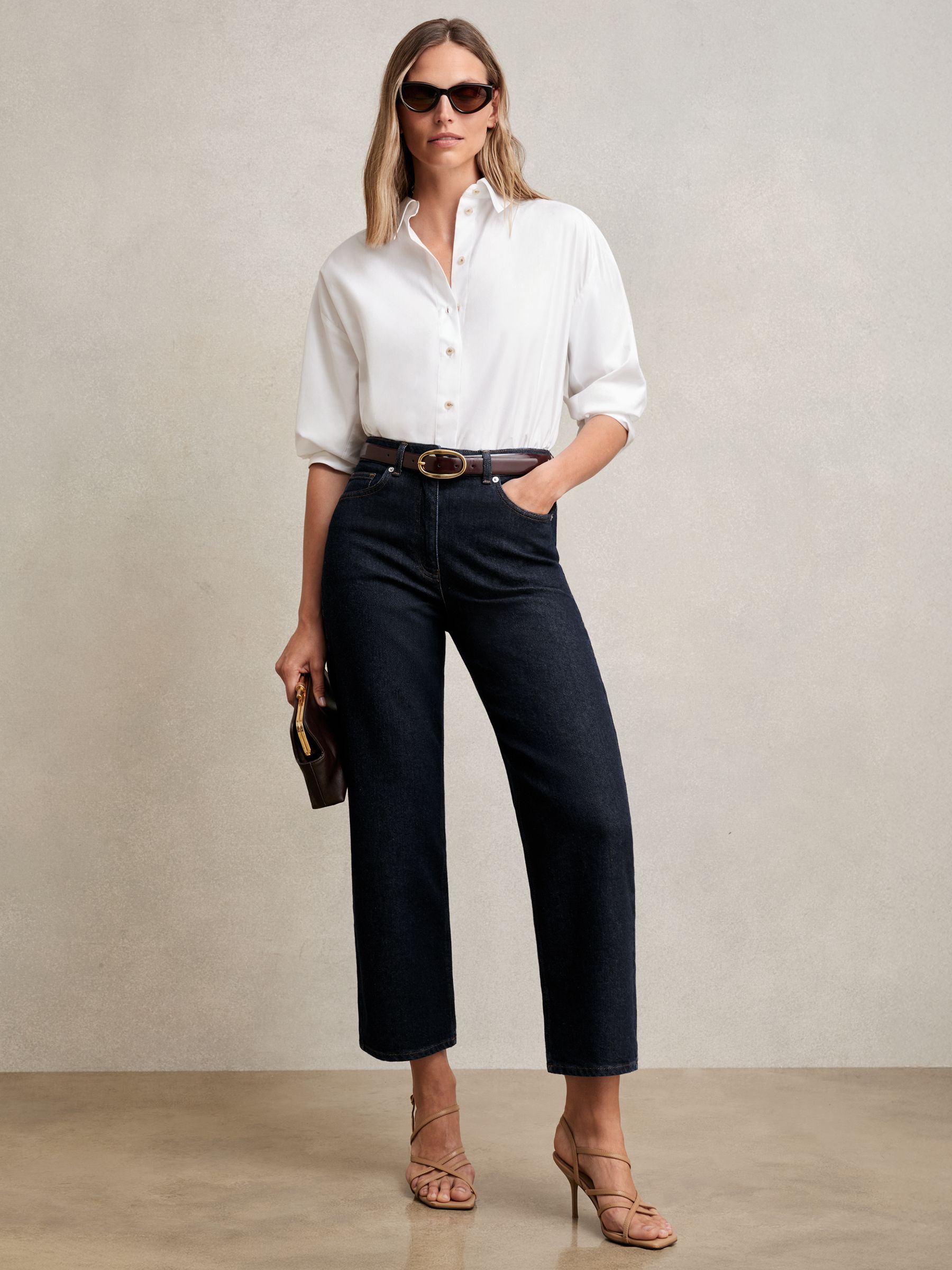 A woman wearing dark blue petite mid-rise straight-leg jeans, paired with a white blouse, sunglasses, and beige heels.