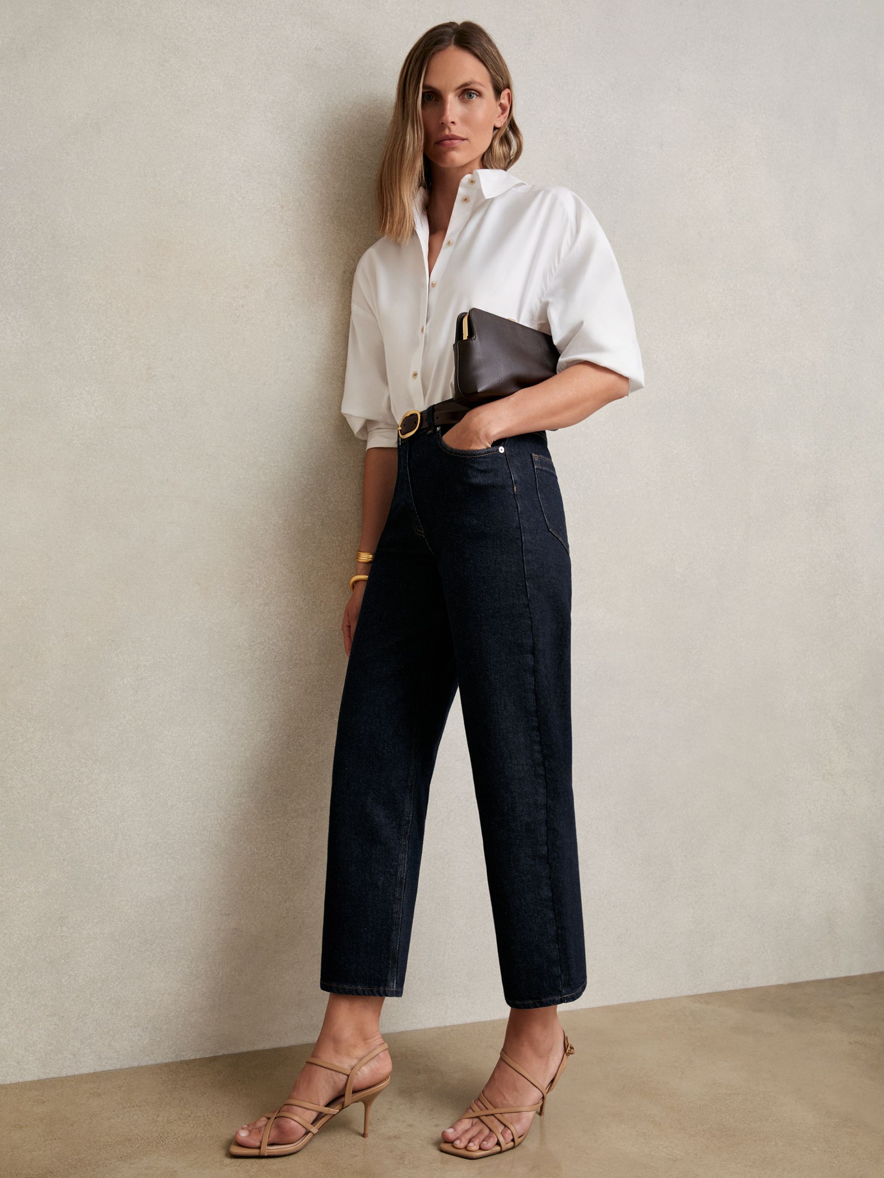 A woman wearing dark blue mid-rise straight-leg jeans with a white blouse, holding a black clutch, and wearing beige high-heeled sandals.