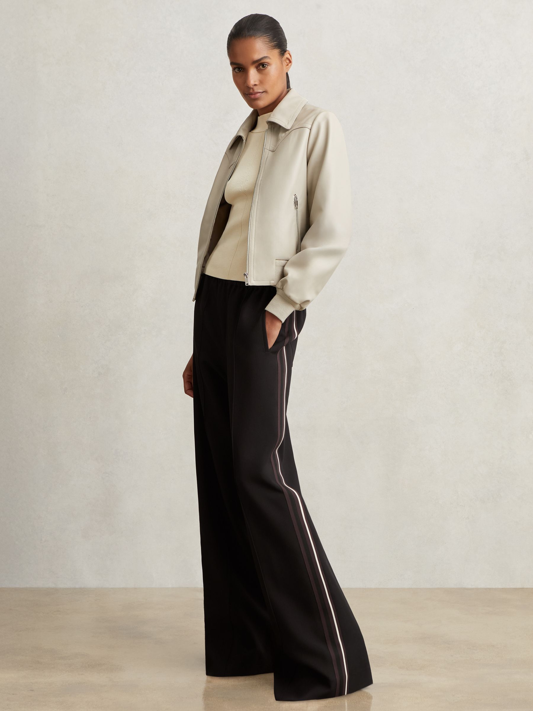 A woman wearing black wide-leg trousers with side stripes, a beige top, and a light-colored jacket, standing against a plain background.