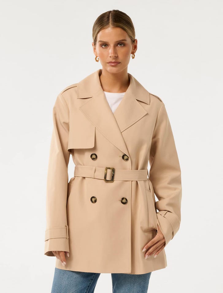 A woman wearing a beige trench coat with notched lapels, double-breasted buttons, a belt with a rectangular buckle, and long sleeves with strap details.