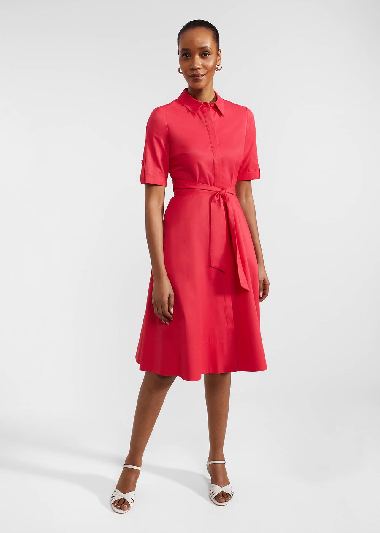 A woman wearing a pink dress with a collar, short sleeves, and a tie belt, standing against a plain background.