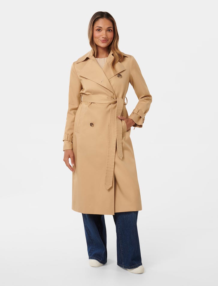 A woman wearing a beige petite soft trench coat with a classic collar, belt, and pockets, standing against a plain background.