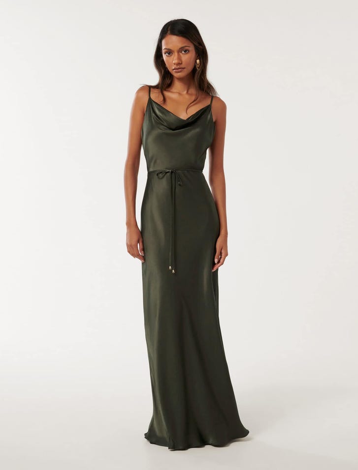 A woman wearing a deep navy satin maxi dress with a cowl neckline and thin straps, standing against a plain background.