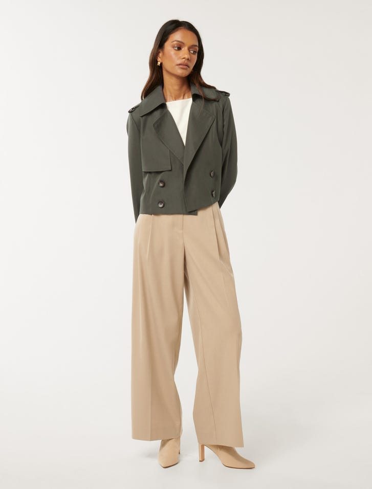 A woman wearing an olive green cropped trench coat with notched lapels, button-up front, and a removable belt, paired with beige wide-leg trousers and beige high-heeled boots.