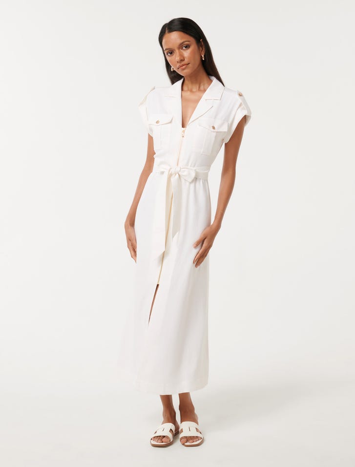 A woman wearing a white midi dress with a notched collar, short sleeves with buttoned cuffs, flap pockets at the bust, an adjustable waist tie, and a zip-through front, standing against a plain background.