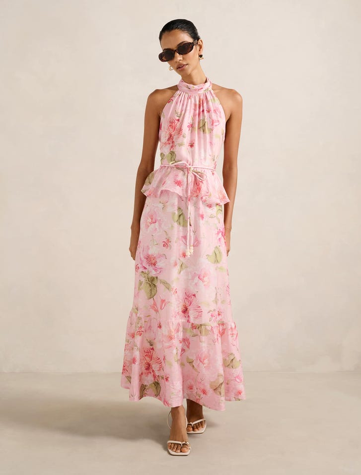 A woman wearing a pink floral print halter neck midi dress with a peplum waist, tiered hem, and bow at the back, paired with sunglasses and heels.