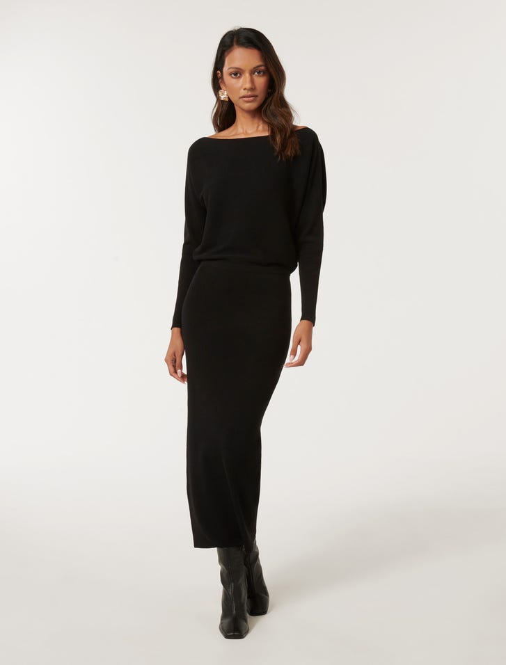 A woman wearing a black off-shoulder midi dress with long sleeves and a fitted silhouette, paired with black boots.