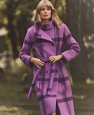 A woman wearing a purple and lavender plaid petite double-breasted coat with a fold-over collar, belt, and long sleeves, standing outdoors.