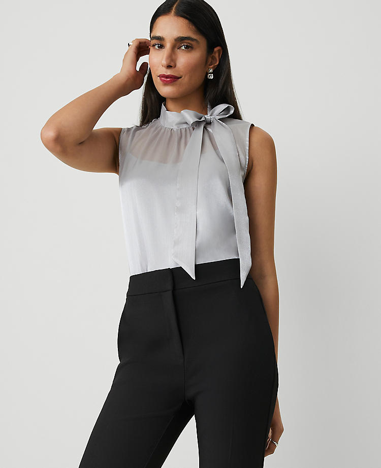 A woman wearing a light grey sleeveless organza blouse with a side-bow tie at the neck, mock neck with ties, shirring below, and a back slit with button closure.