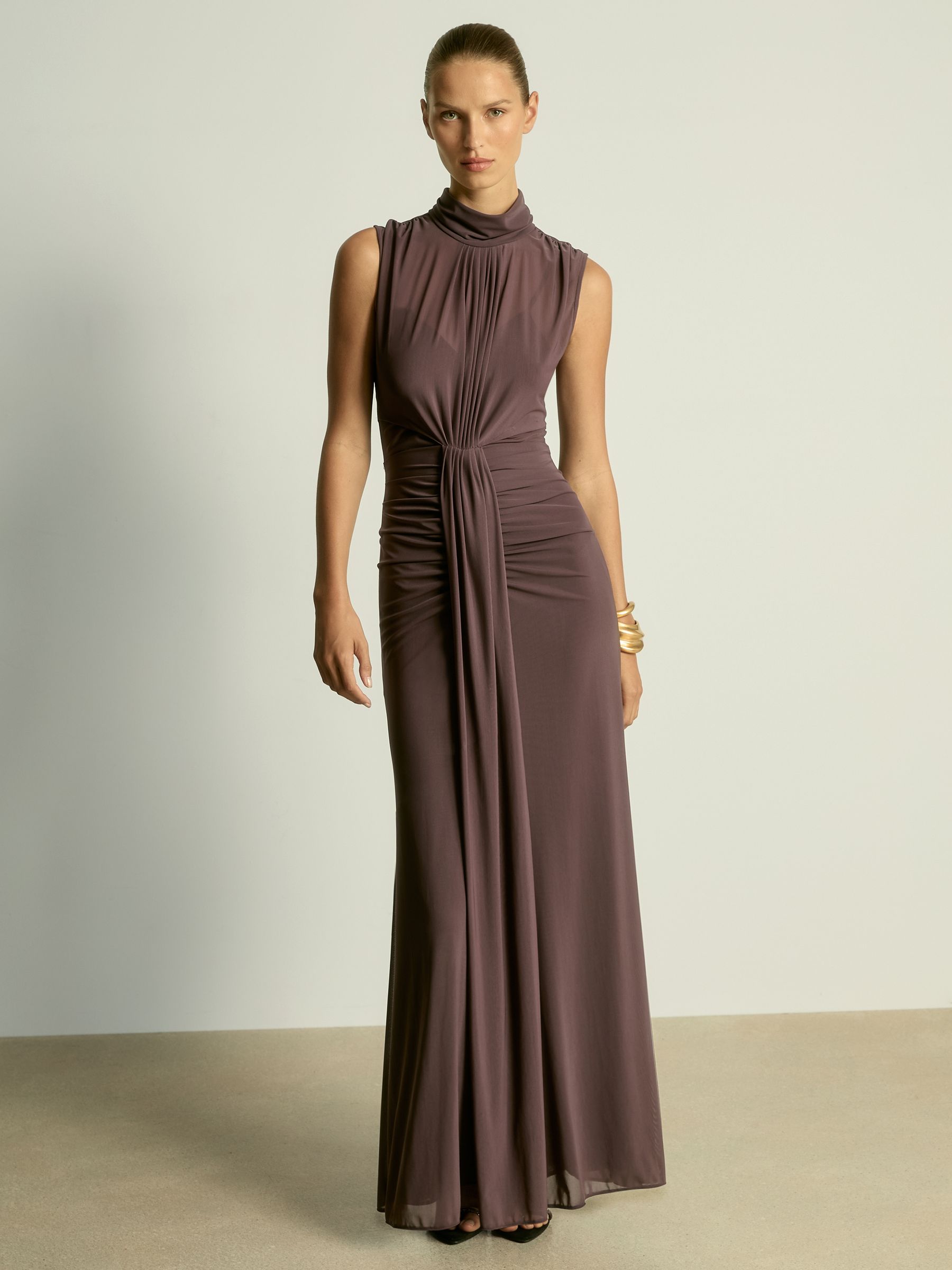 A woman wearing a taupe sheer ruched maxi dress with a high neckline and sleeveless design.