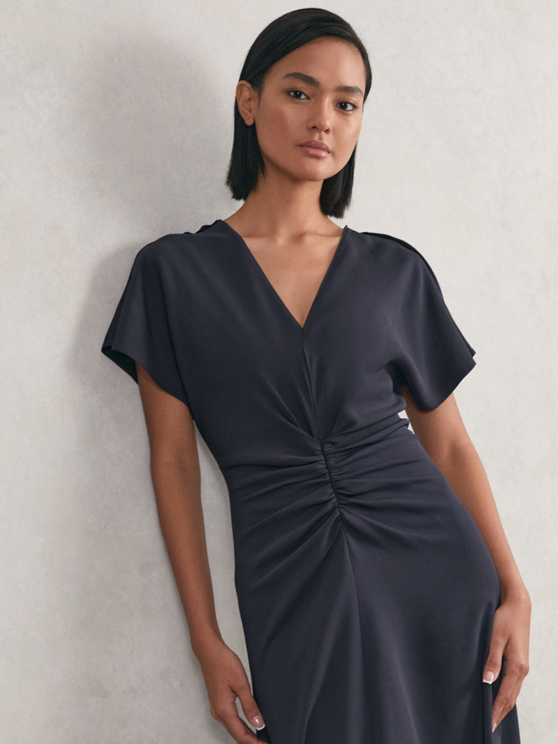 A woman wearing a navy midi dress with ruched front, flutter sleeves, V-neckline, and a fitted silhouette.