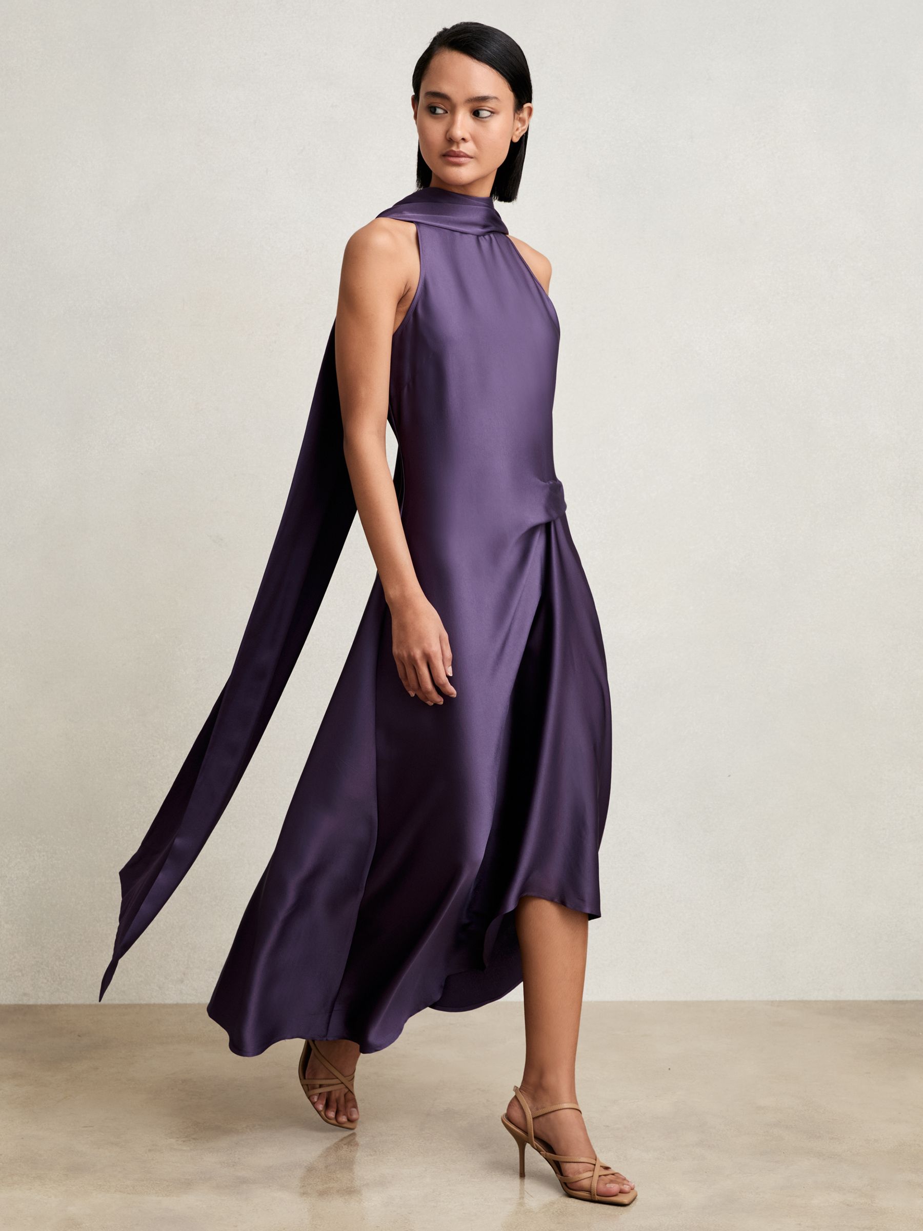 A woman wearing a purple satin midi dress with a cape back and tuck detail at the waist.