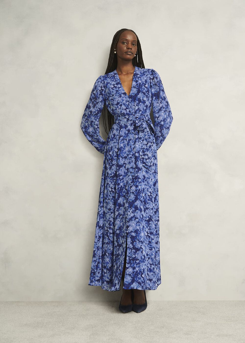 A woman wearing a long blue floral dress with a V-neckline, long blouson sleeves, a waist tie, and side pockets.