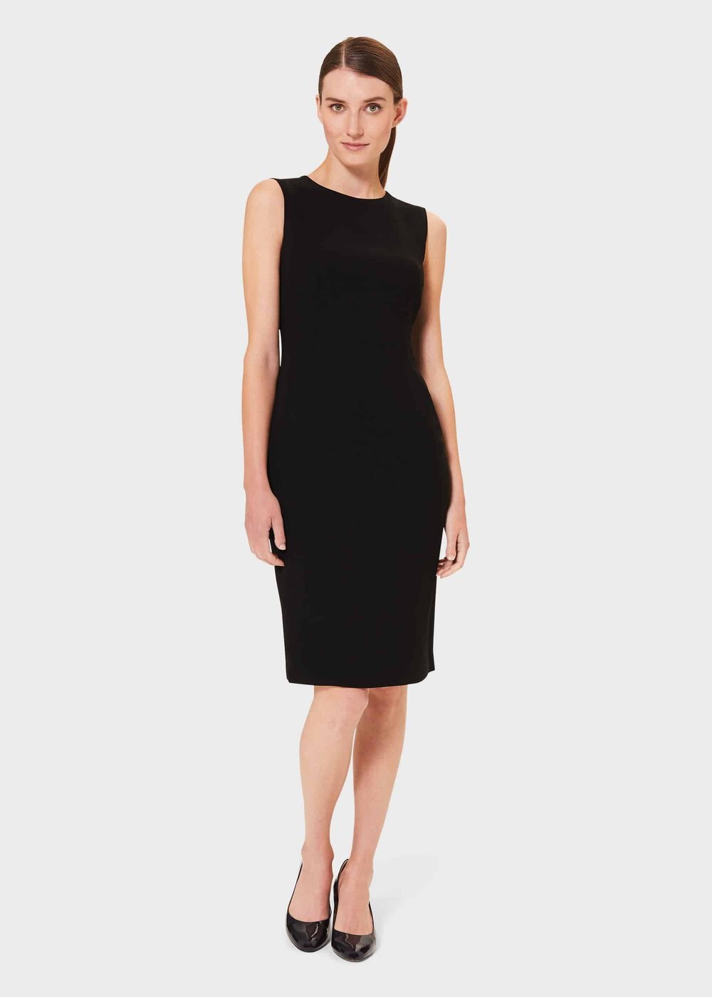 A woman wearing a sleeveless black dress with a crew neckline, fitted silhouette, and midi length, paired with black heels.