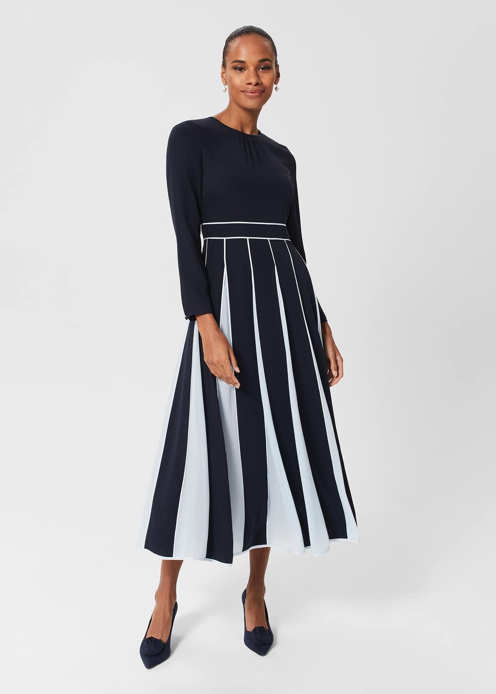 A woman wearing a navy blue fit and flare dress with white contrast godet pleats, long sleeves, and a crew neckline, standing against a plain background.