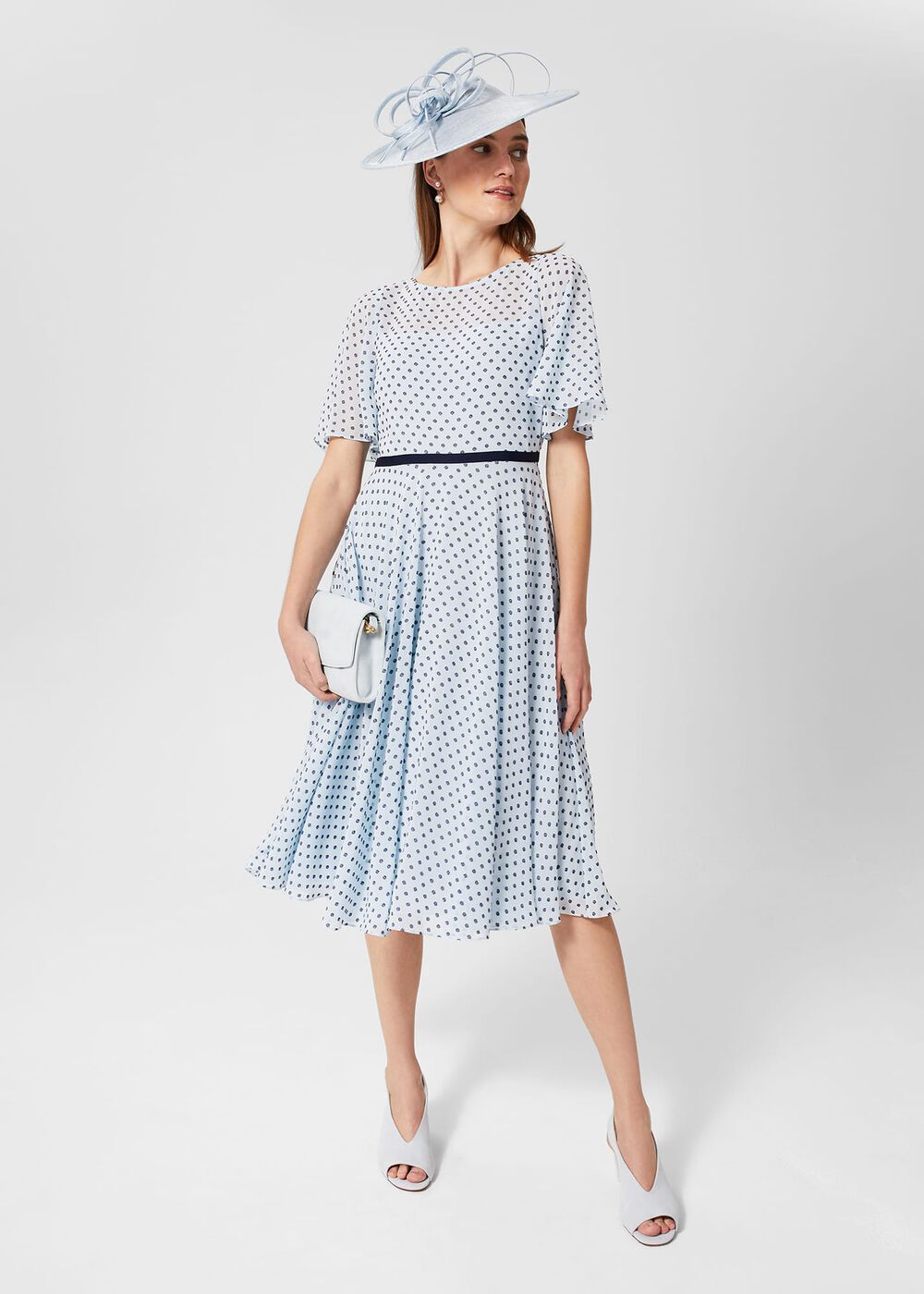 A woman wearing a white dress with navy polka dots, flared skirt, and fluted sleeves, accessorized with a white clutch and a large white hat with decorative elements.