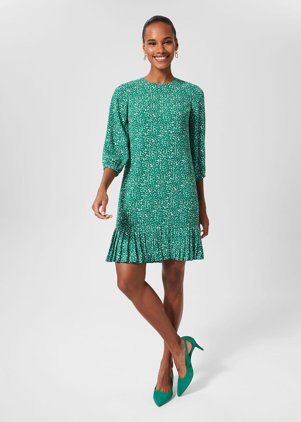 A woman wearing a green dress with a white spot print, featuring a crew neckline, three-quarter-length sleeves, and a pleated hem, paired with green high heels.