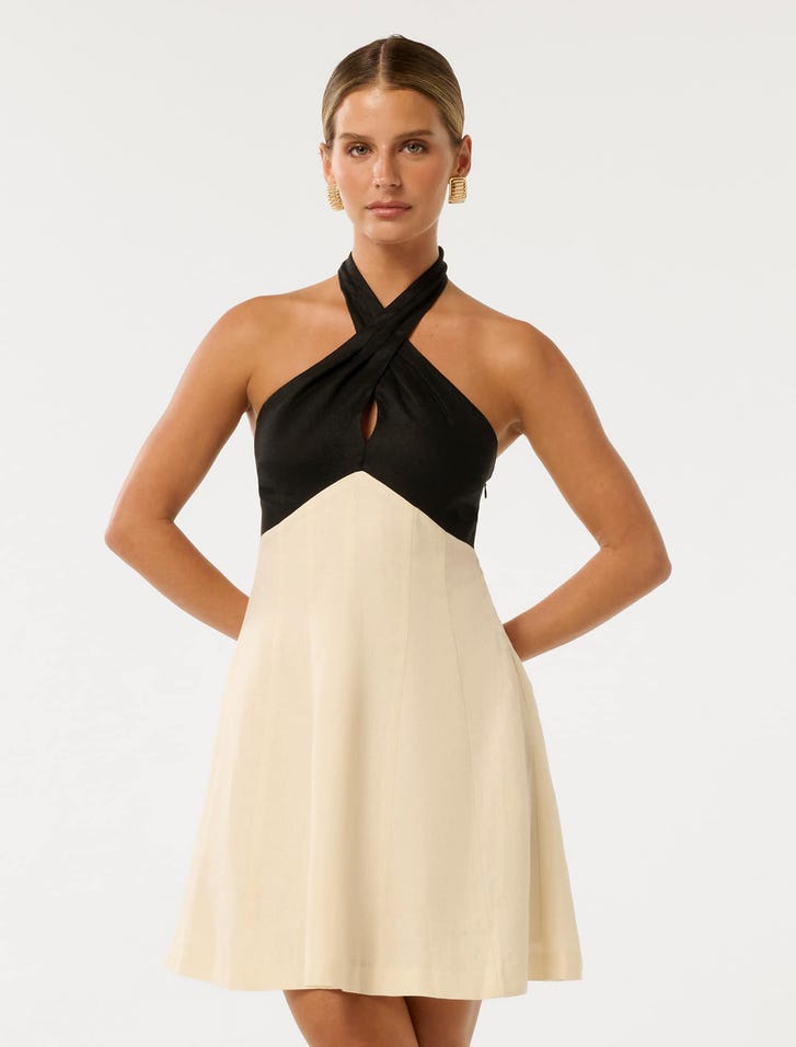 A woman wearing a petite black and cream halter neck mini dress with a crossover neckline, keyhole detail, and high-waisted A-line skirt.