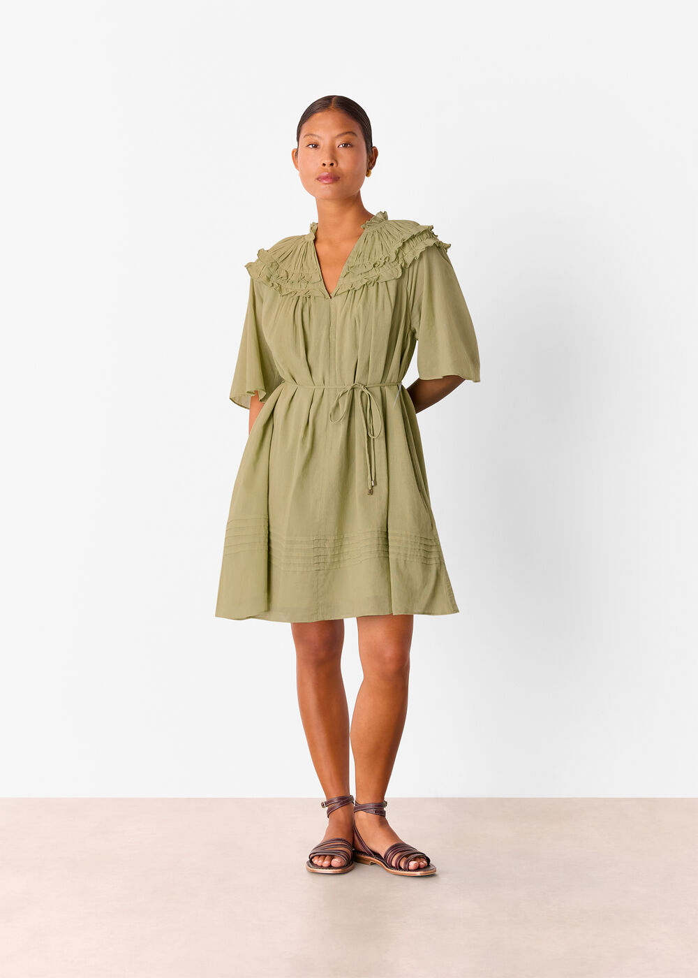 A woman wearing a khaki mini dress with frill details at the shoulders and chest, a tie belt at the waist, and a relaxed fit.