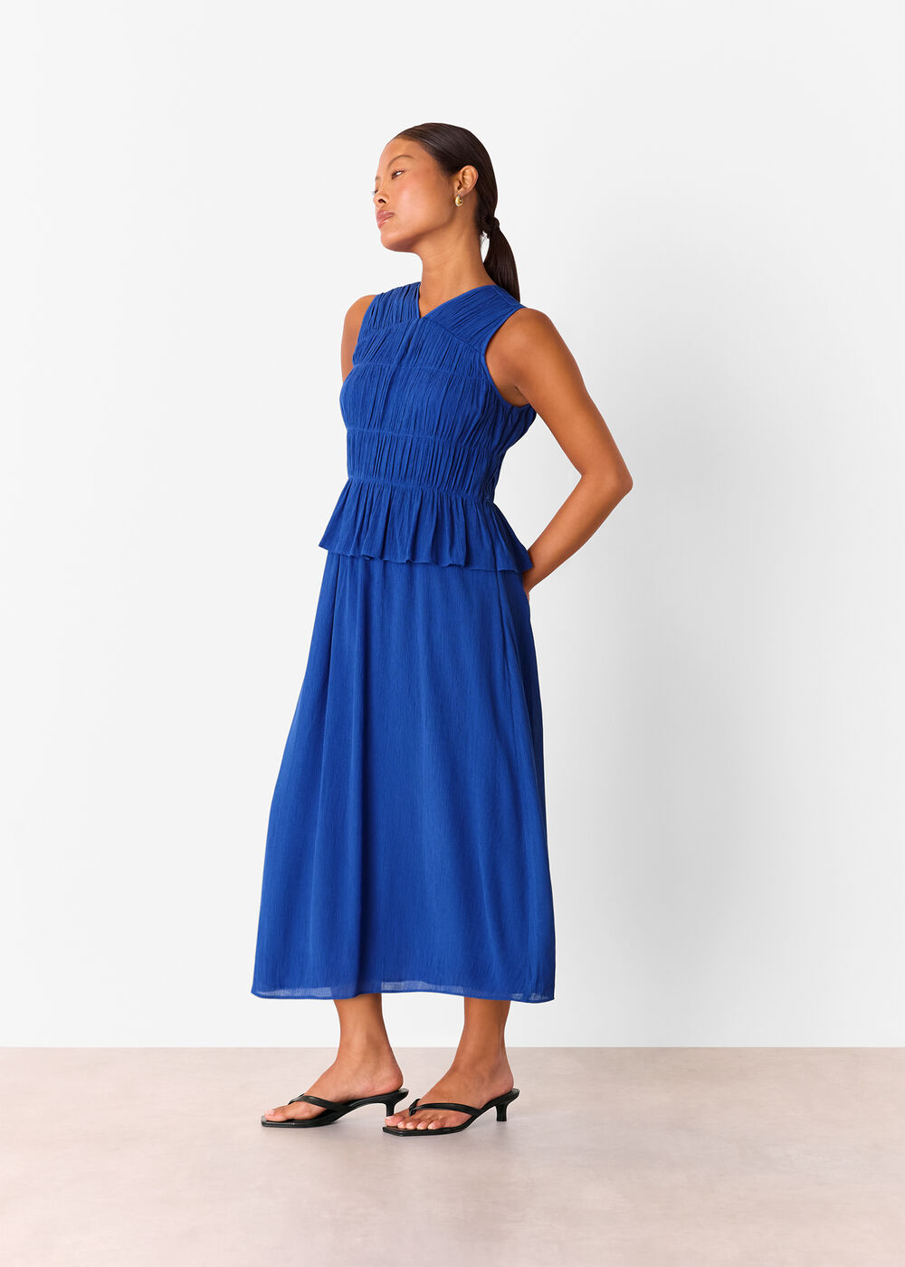 A woman wearing a cobalt blue midi dress with shirring details on the bodice, a V-neckline, a fitted waist with ruffles, and a flowing skirt, paired with black heeled sandals.