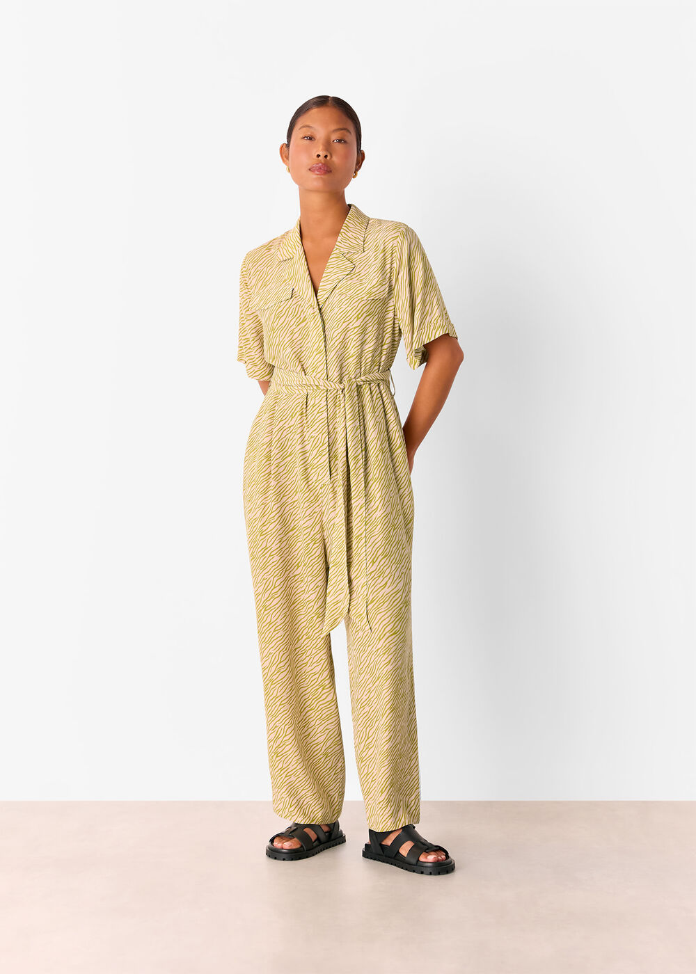 A woman wearing a yellow-green jumpsuit with a tiger flame pattern, collar, V-neckline, front pockets, and a belt, paired with black sandals.