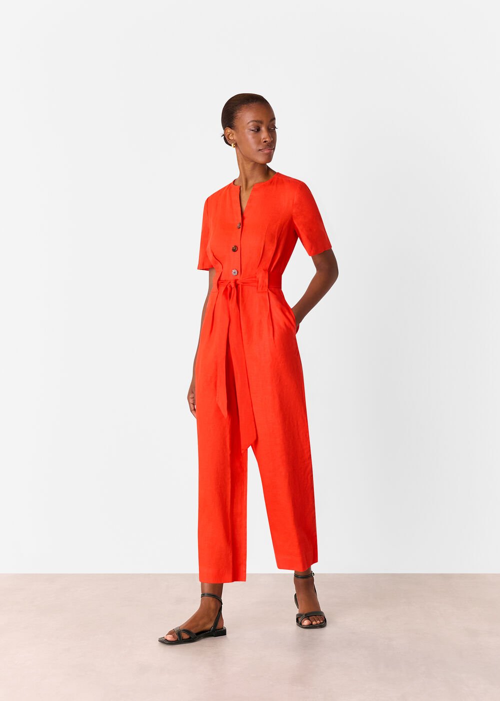 A woman wearing a bright red linen jumpsuit with short sleeves, a round neckline, a belt at the waist, and button details in front.