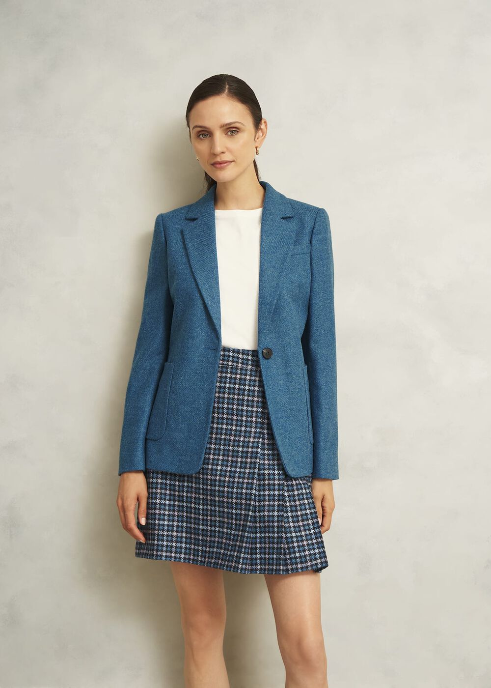 A woman wearing a blue tailored wool jacket with notch lapels and a melton undercollar, paired with a white top and a checkered skirt.