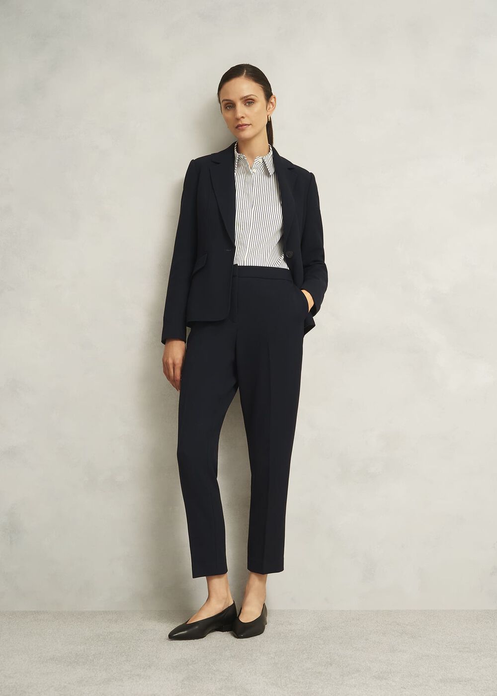 A woman wearing navy blue slim fit trousers with a matching blazer, a white and black striped shirt, and black shoes, standing against a plain light background.