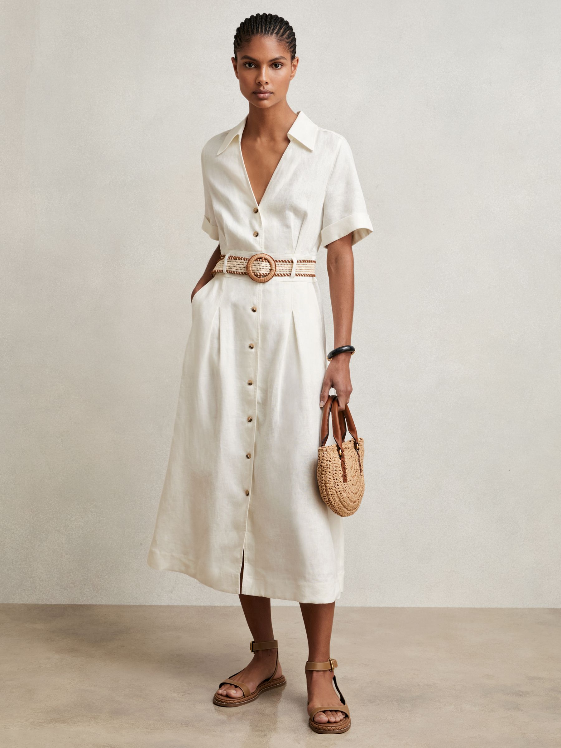 A woman wearing a white linen midi shirt dress with a collar, V-neckline, front buttons, a woven raffia belt, turn-up cuffs, and front pleats.