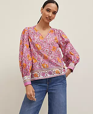 A woman wearing a pink floral V-neck blouse with long puffed sleeves and a shirttail hem.