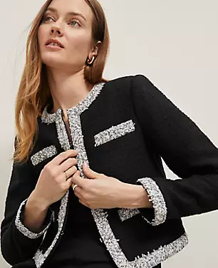 A woman wearing a black petite cropped tweed jacket with white trim, round neck, long sleeves, and front pockets.